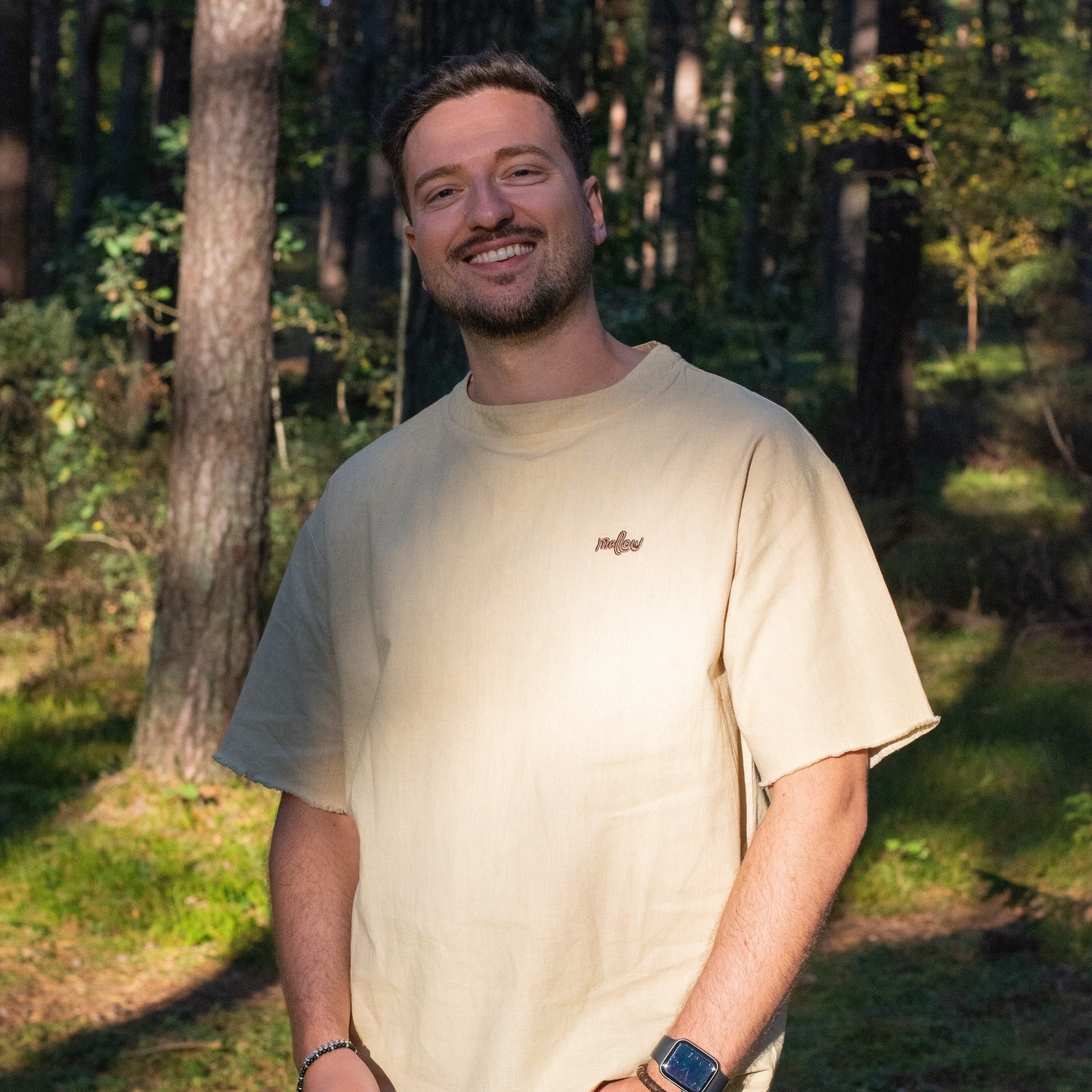 A man standing in a forest setting, wearing a creamy beige hemp tee with short sleeves, jeans, and a watch on his left wrist.