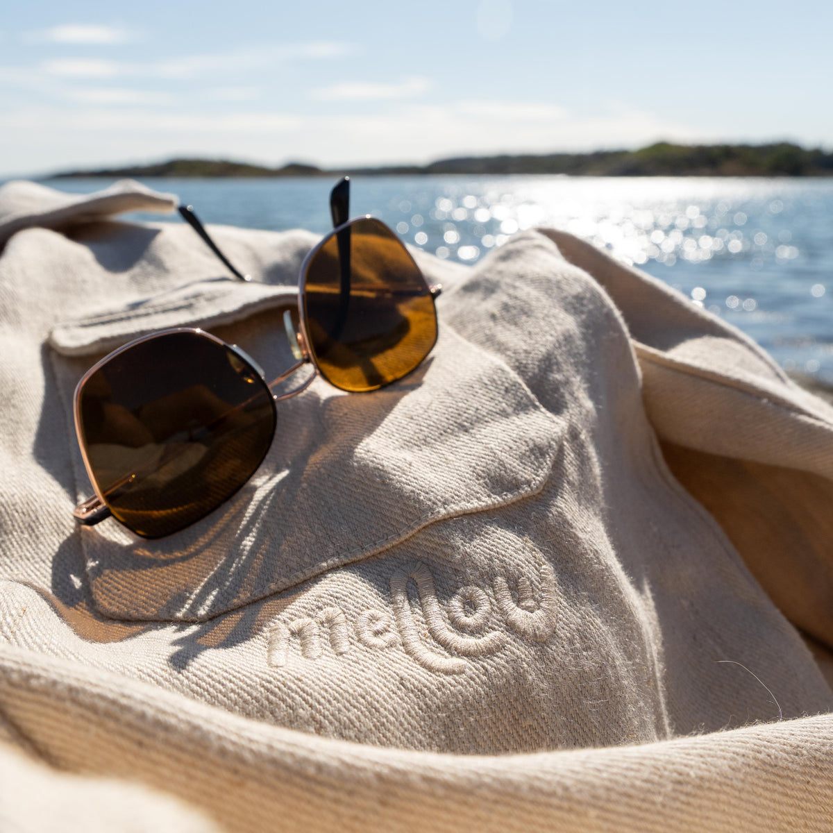 A pair of brown aviator sunglasses resting on a beige woven hemp jacket with the brand name 'mellow' embroidered on it, set against a blurred background of water and sky.