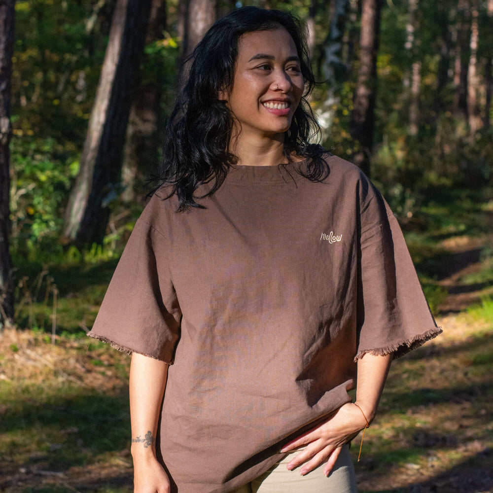 A person standing in a forest setting, wearing an earthy brown oversized hemp tee with short sleeves.
