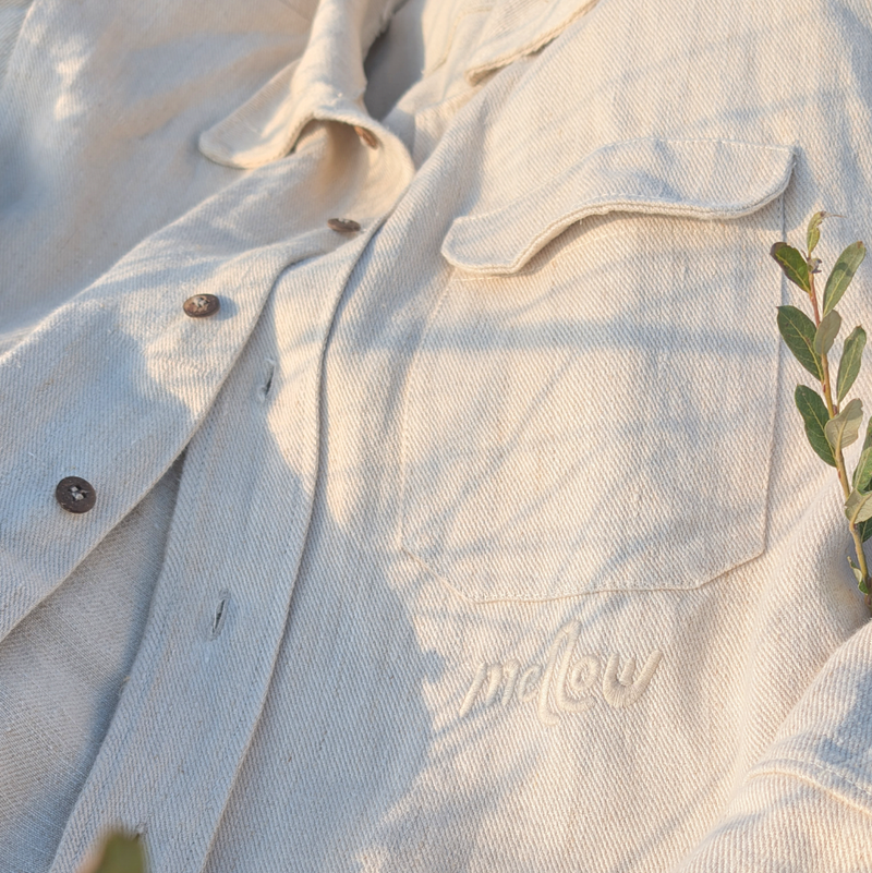 A light beige hemp button-up shirt with 'mellow' embroidered on the chest, laid flat on a grassy background with natural sunlight and shadows.