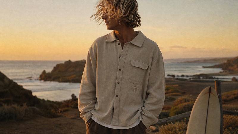 Man standing on a beach at sunset with a surfboard, wearing a beige Hemp overshirt.