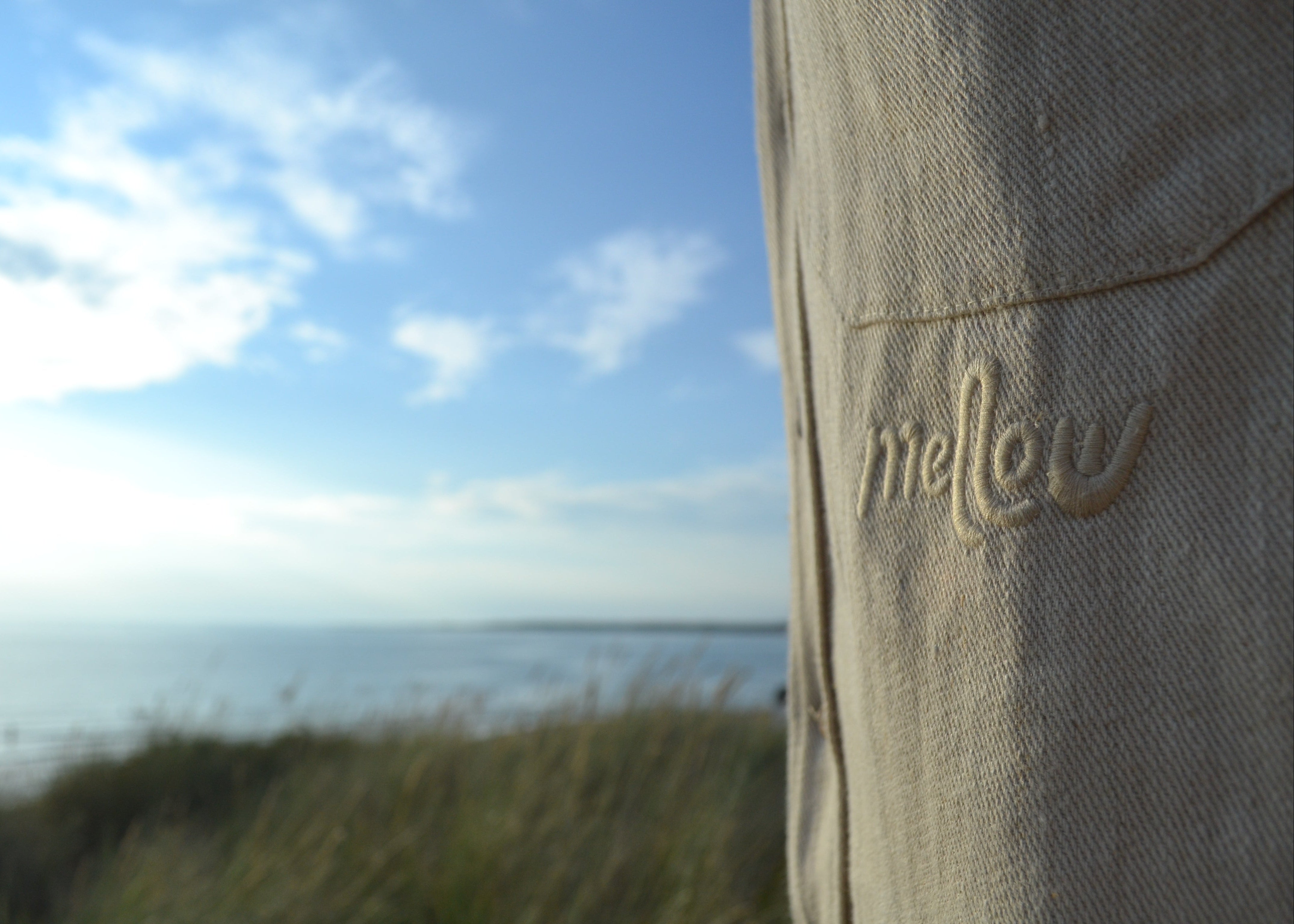 Close-up of a beige hemp garment with the embroidered brand name 'mellow' on the pocket, set against a blurred outdoor background of blue sky, sea, and grass.