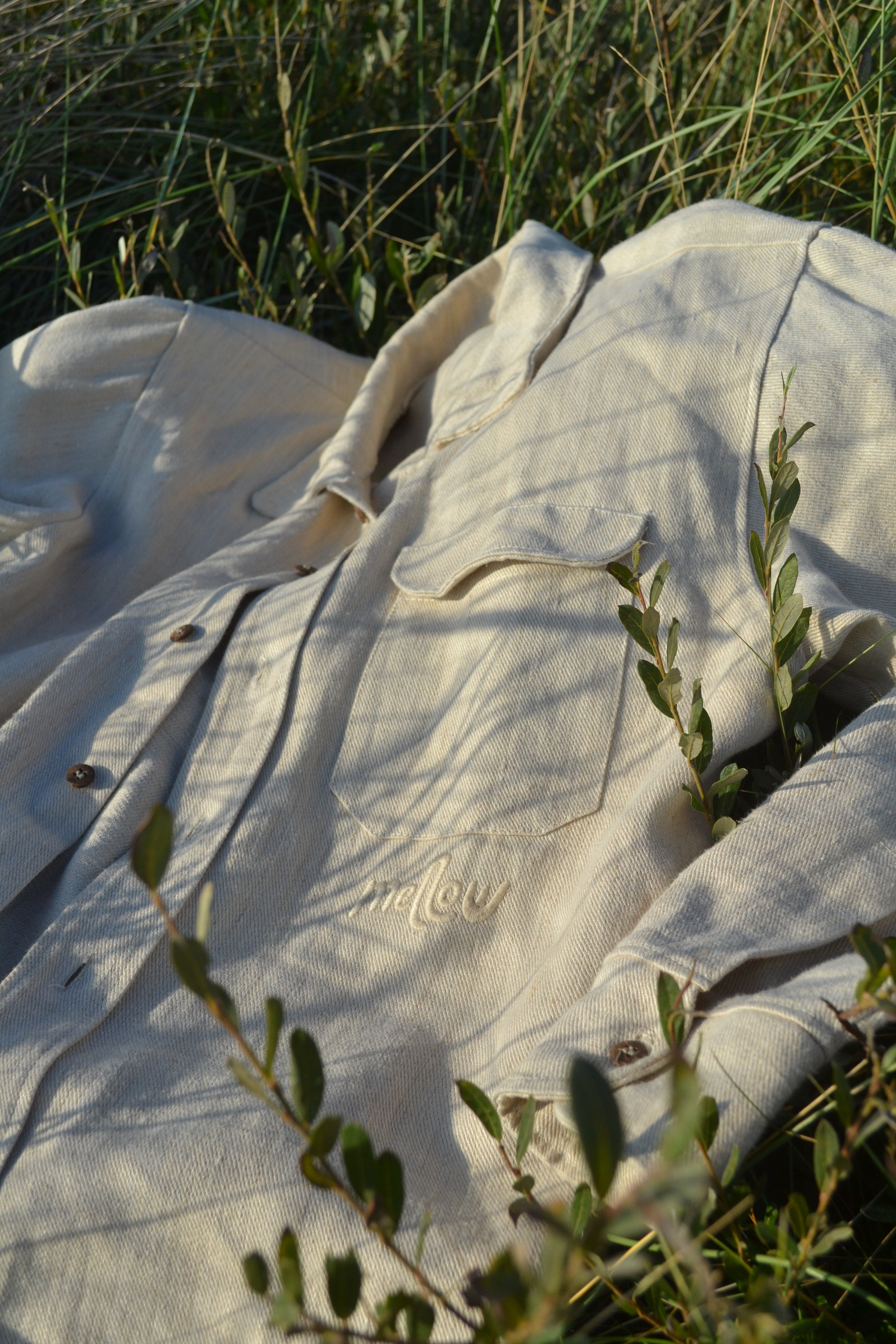 A light beige hemp button-up shirt with a tonal embroidered logo, laid out in a field of grass with natural foliage in the foreground.