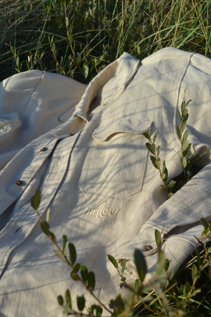 A light beige hemp button-up shirt with a tonal embroidered logo, laid out in a field of grass with natural foliage in the foreground.
