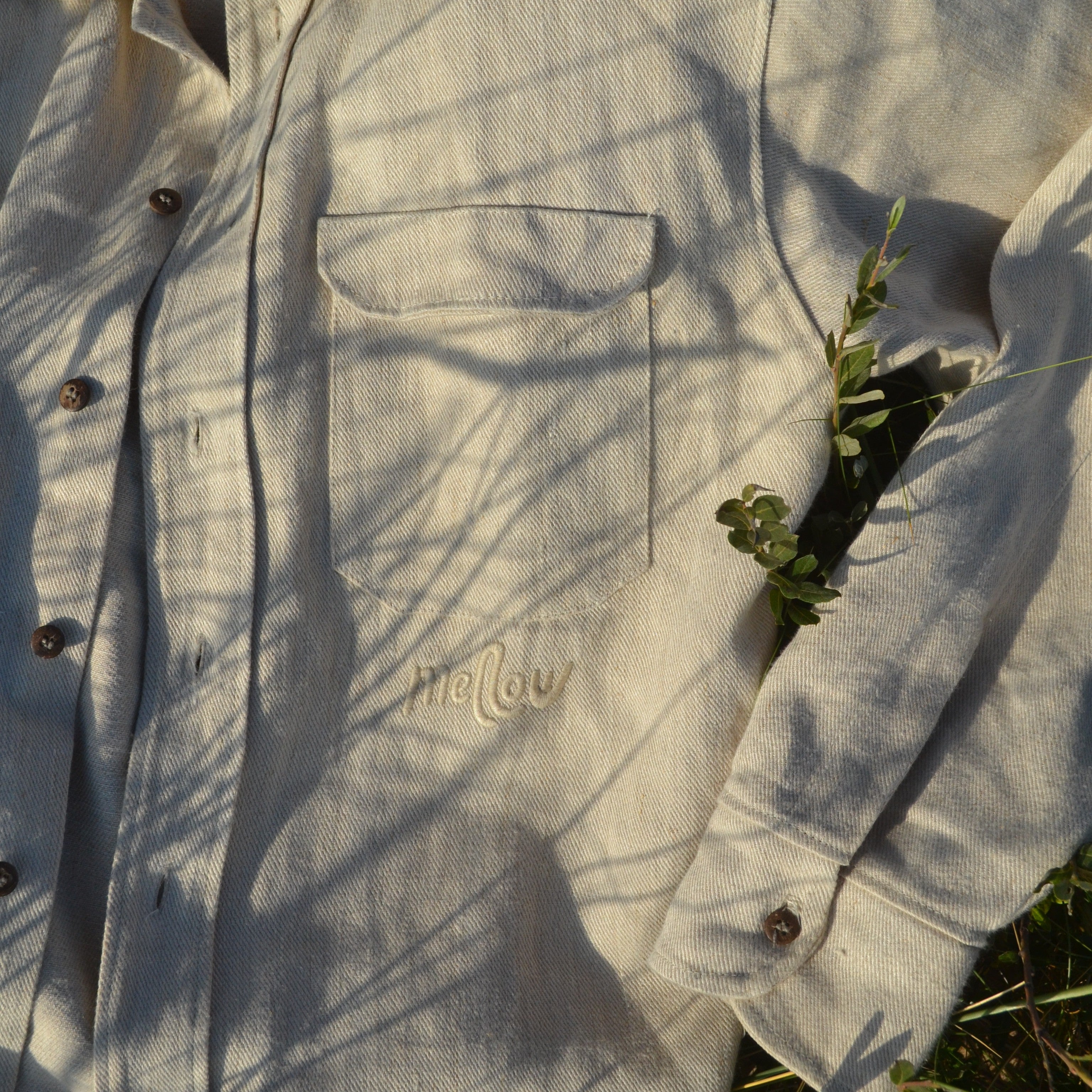 A close-up of a light beige hemp button-up shirt with a chest pocket and embroidered 'Mellow' script on the front, styled outdoors with a small green plant tucked into the pocket under dappled sunlight.