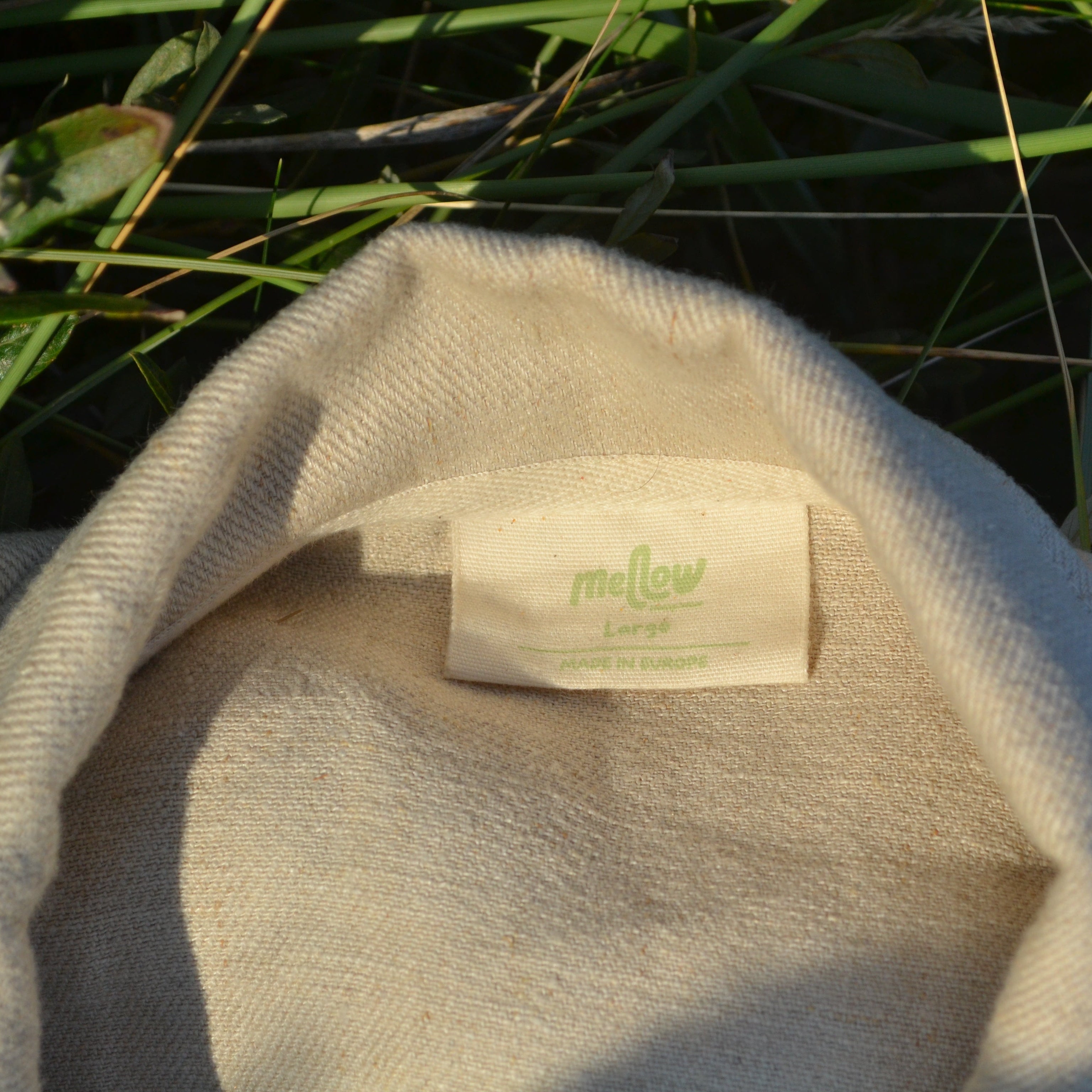 Close-up of a cream-colored hemp garment with a visible sewn-in label reading 'mellow', photographed outdoors against green grass in warm sunlight.