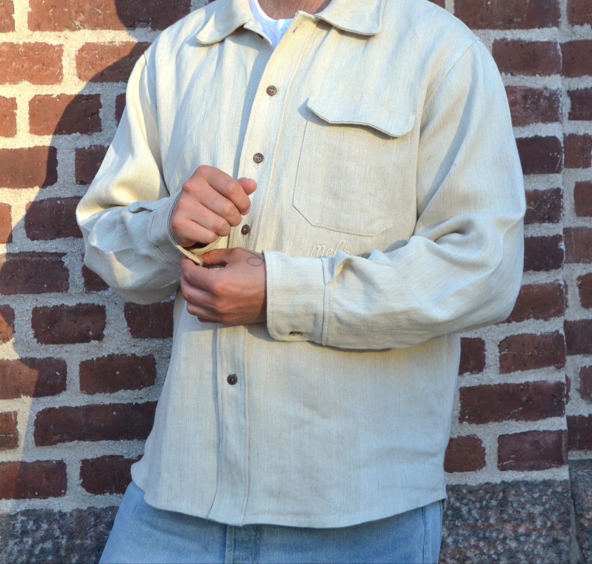 A man wearing a light hemp linen button-up shirt over a white undershirt, standing against a red-brown brick wall.