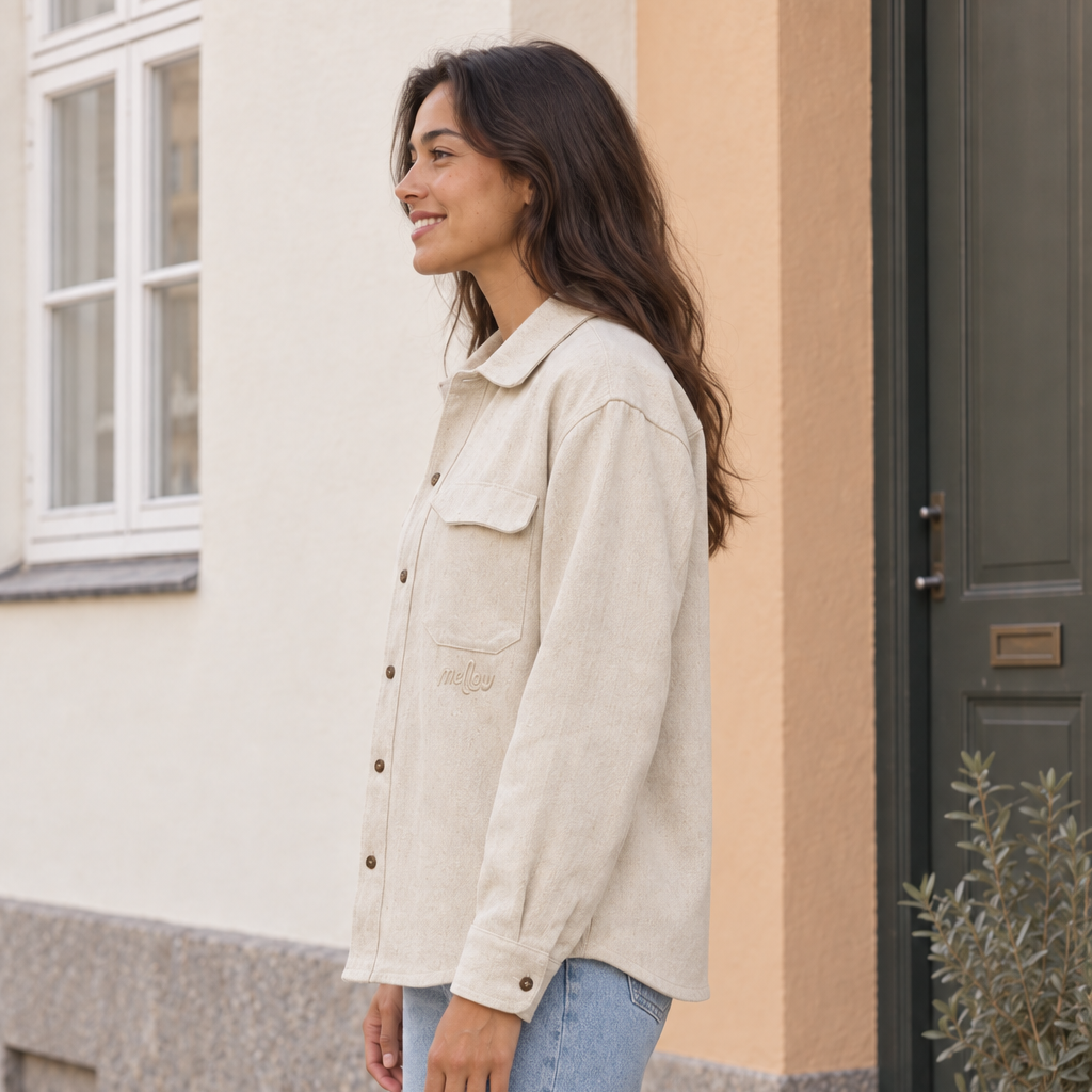 A woman wearing a light beige hemp button-up shirt and light blue jeans stands on a cobblestone street in front of a building with a beige wall and a dark green door.