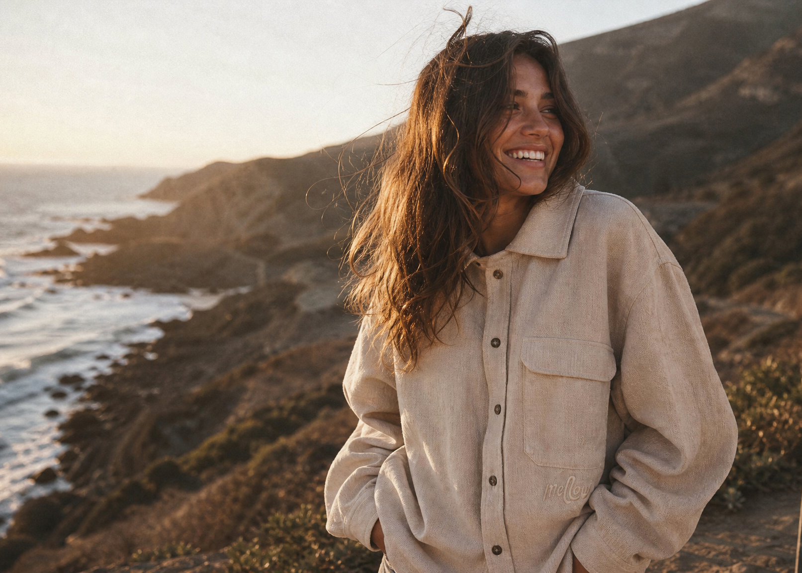 Woman standing by the ocean with a hemp jacket and skateboard, smiling at sunset.